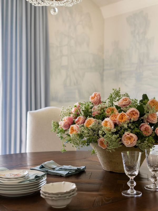 A large arrangement of peach garden roses centers a round wood dining table beneath a crystal chandelier in a mural-lined dining room.