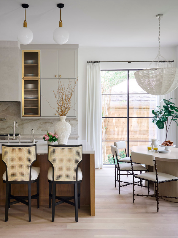A bright kitchen with cane-backed counter stools, a sculptural vase, and a round breakfast nook near floor-to-ceiling windows.