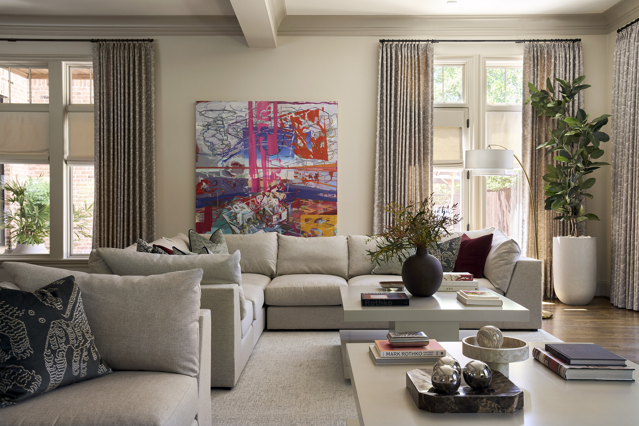 Light-filled Hunter’s Creek living room with neutral sectional sofas, textured gray drapery, modern white coffee tables, greenery, and burgundy accents.