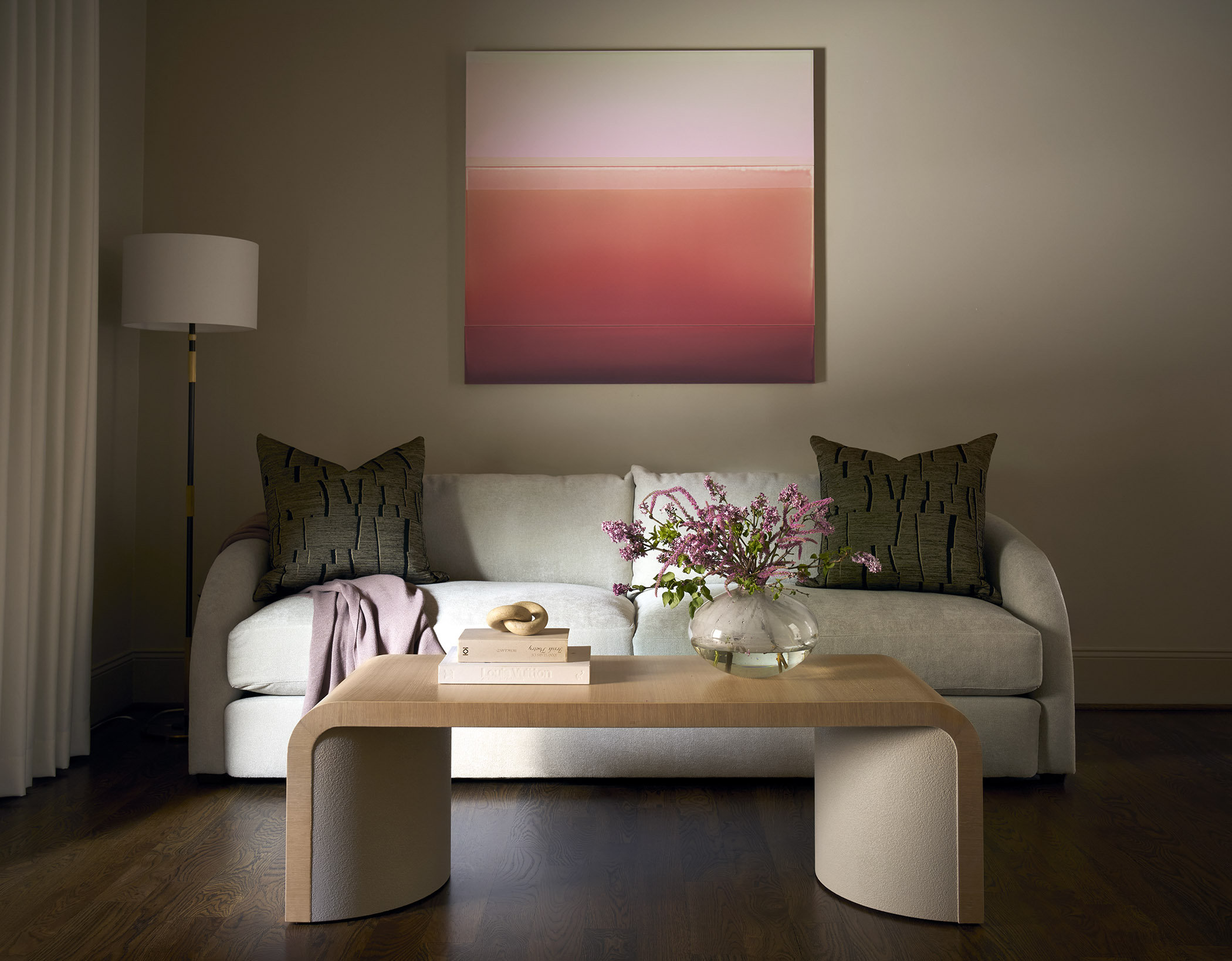 Intimate sitting area with a curved cream sofa, wood and upholstered coffee table, pink abstract artwork, dark wood floors, and fresh flowers.