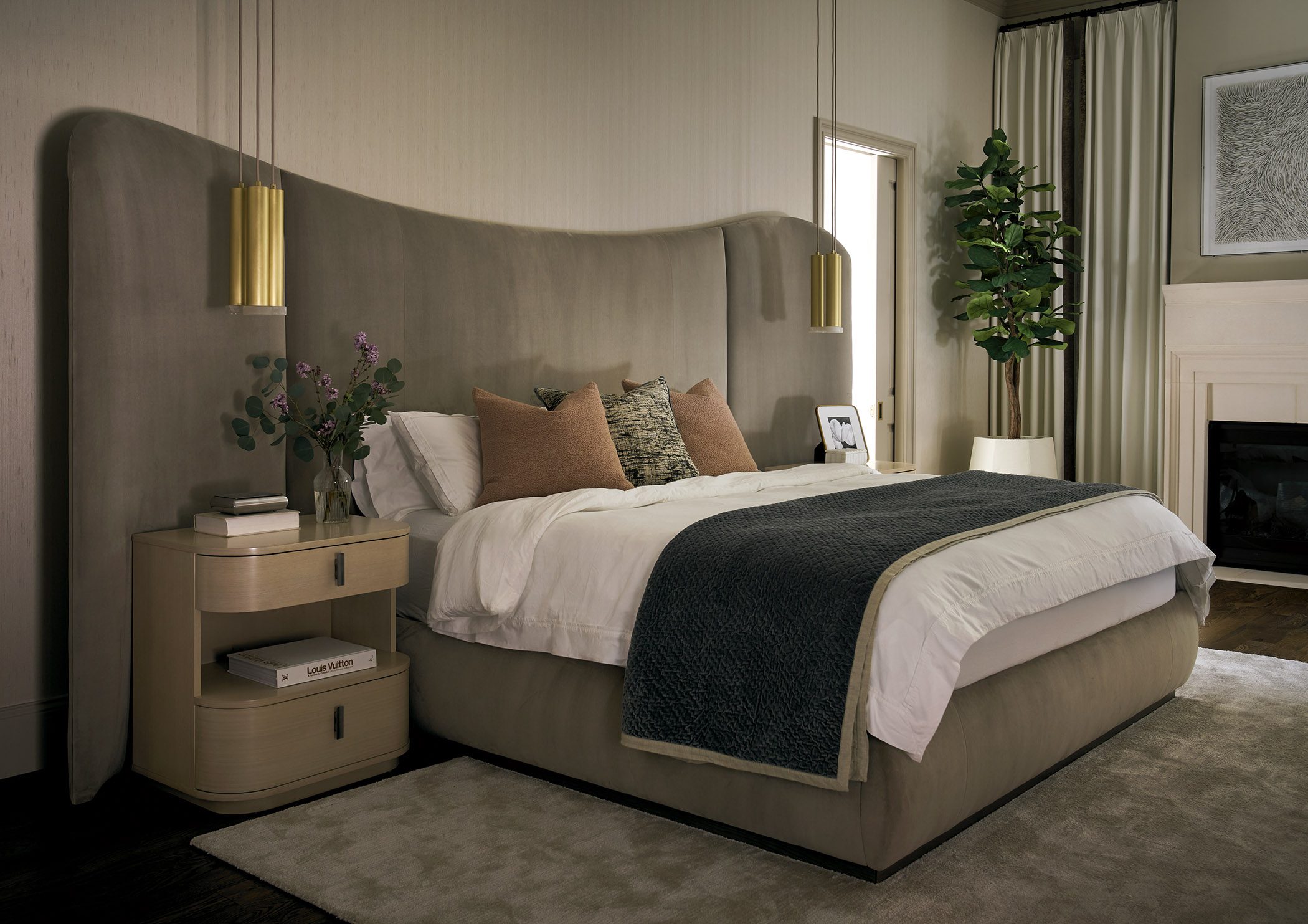 Serene primary bedroom with a sweeping upholstered headboard, brass pendant lights, neutral bedding, dark textured throw, fireplace, and tall indoor tree.