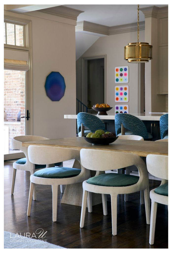 Bright breakfast room with white upholstered chairs, deep teal seat cushions, a warm wood table, blue patterned counter stools, colorful art, and a brass pendant.