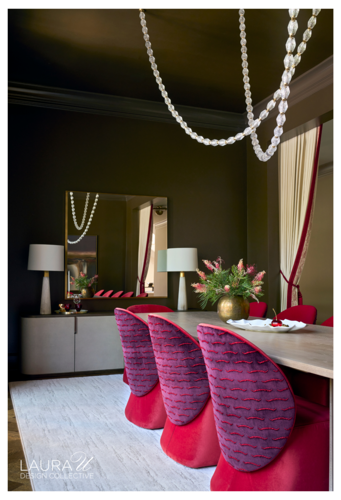 Long view of the olive dining room with cherry-red sculptural chairs, a pale table and rug, looping glass chandelier, mirrored sideboard, and red-trimmed drapery.