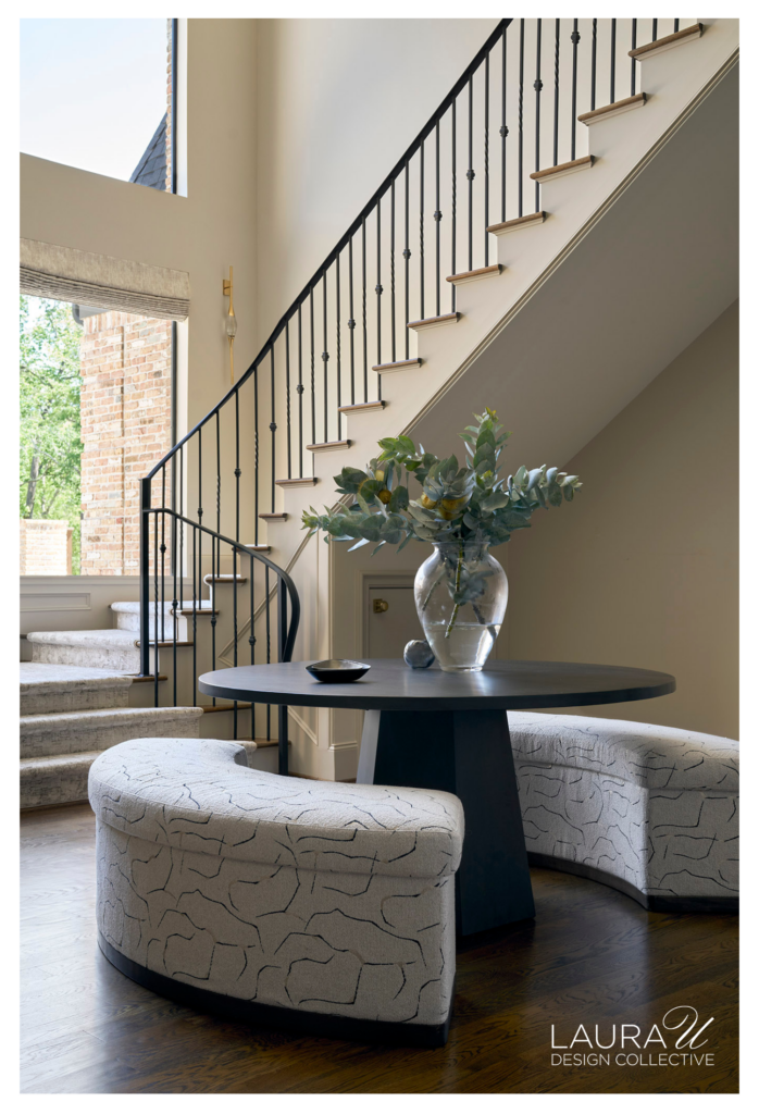 Light-filled stair hall with black iron railing, dark wood floors, a round black table, and curved upholstered benches beneath the sweeping staircase.