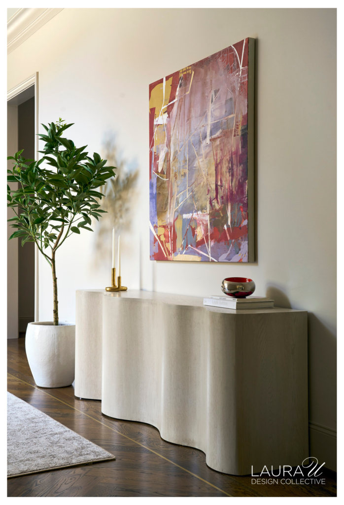 Hallway vignette with a sculptural white oak console, colorful abstract artwork, brass candlesticks, a silver bowl, and a tall potted tree.