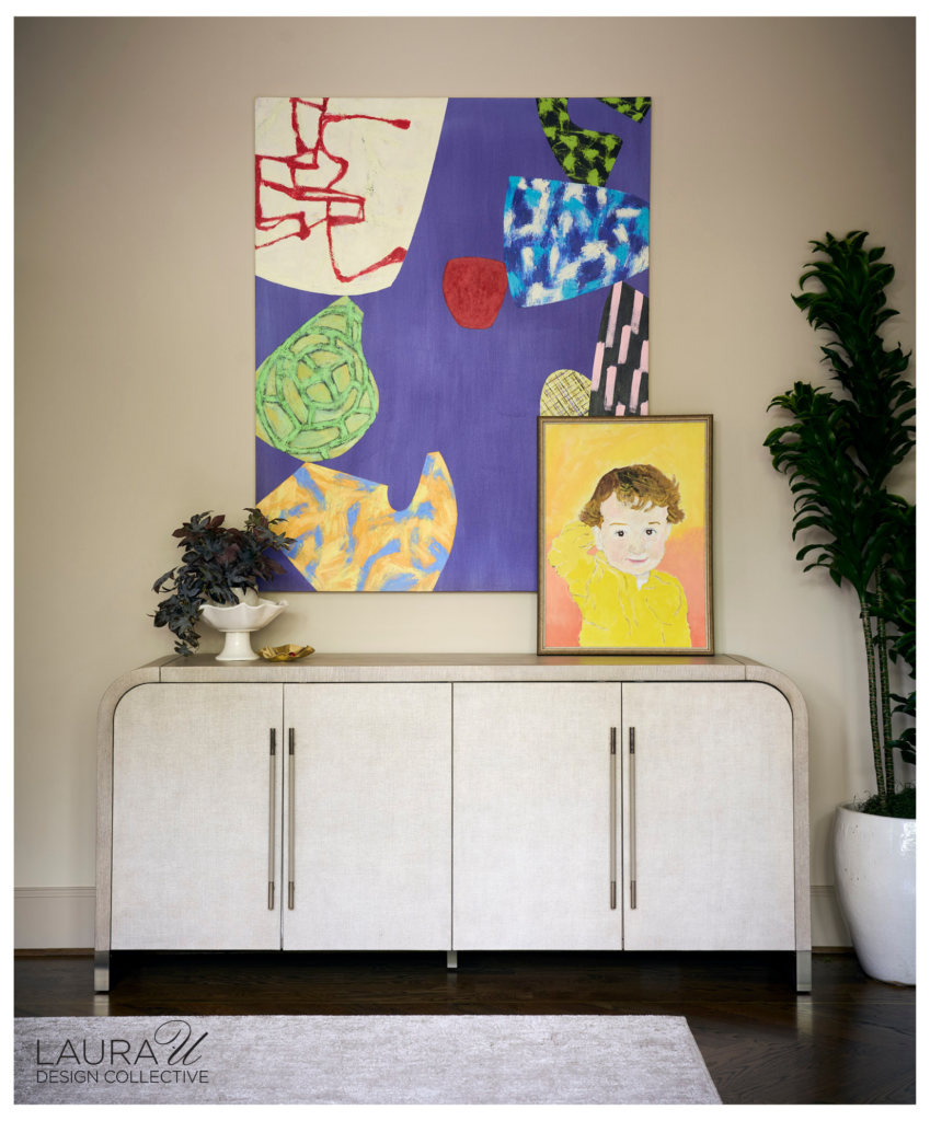 Rounded cream buffet topped with a colorful child portrait and decorative objects beneath a large purple abstract painting, flanked by a tall green plant.