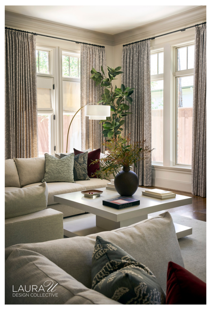 Spacious living room with pale sectional seating, colorful abstract art, textured gray drapery, white cocktail tables, patterned pillows, and lush greenery.