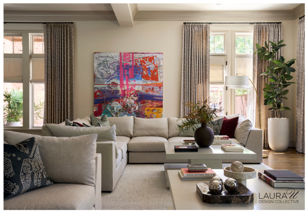 Spacious living room with pale sectional seating, colorful abstract art, textured gray drapery, white cocktail tables, patterned pillows, and lush greenery.