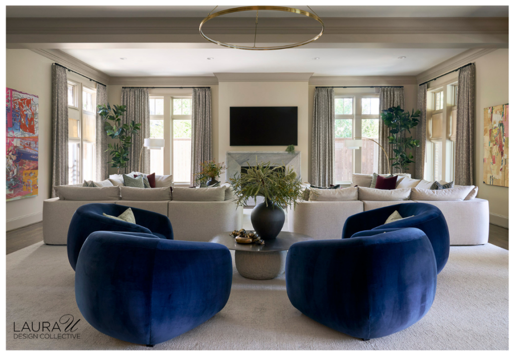 Four cobalt velvet lounge chairs surround a sculptural metallic cocktail table in front of pale sectional sofas, tall windows, greenery, and colorful abstract art.