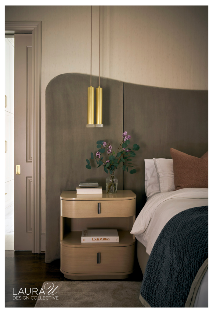 Primary bedroom with an oversized curved taupe headboard, brass pendant lights, rounded nightstands, white bedding, a dark blue throw, fireplace, and tall potted tree.