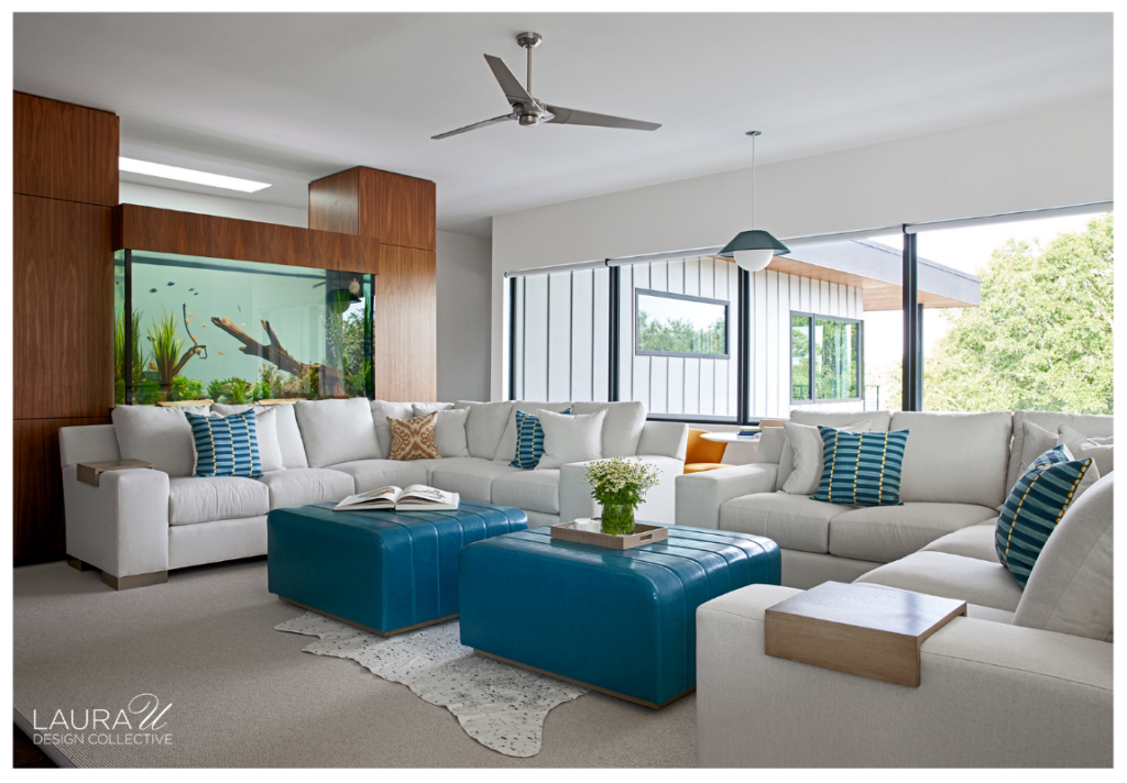 Braeswood family room featuring expansive white sectional sofas, teal leather ottomans, striped accent pillows, floor-to-ceiling windows, and a large aquarium integrated into custom walnut cabinetry.
