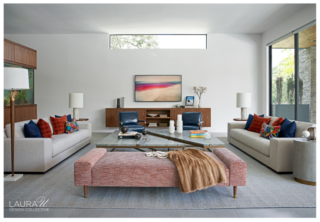 Braeswood living room with two light gray sofas, colorful accent pillows, blue leather chairs, layered stone coffee tables, and a coral upholstered bench beneath a contemporary abstract painting.