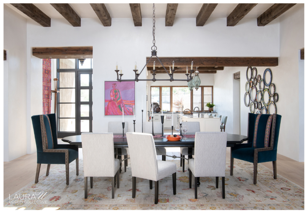 Camino Haciendas dining room featuring a dark wood dining table, teal and cream upholstered chairs, a wrought-iron chandelier, rustic ceiling beams, and a colorful gallery of decorative mirrors.