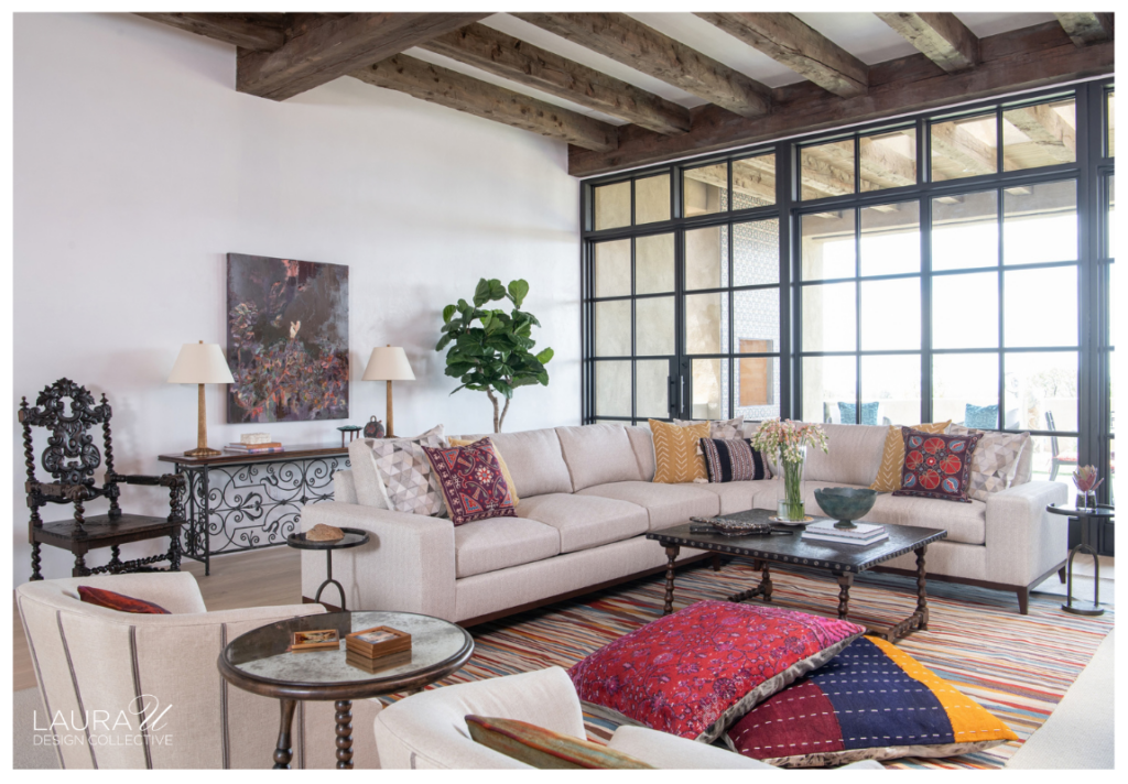 Camino Haciendas living room featuring exposed rustic wood beams, a large cream sectional sofa, colorful patterned pillows, an antique-style coffee table, and a vibrant striped area rug.