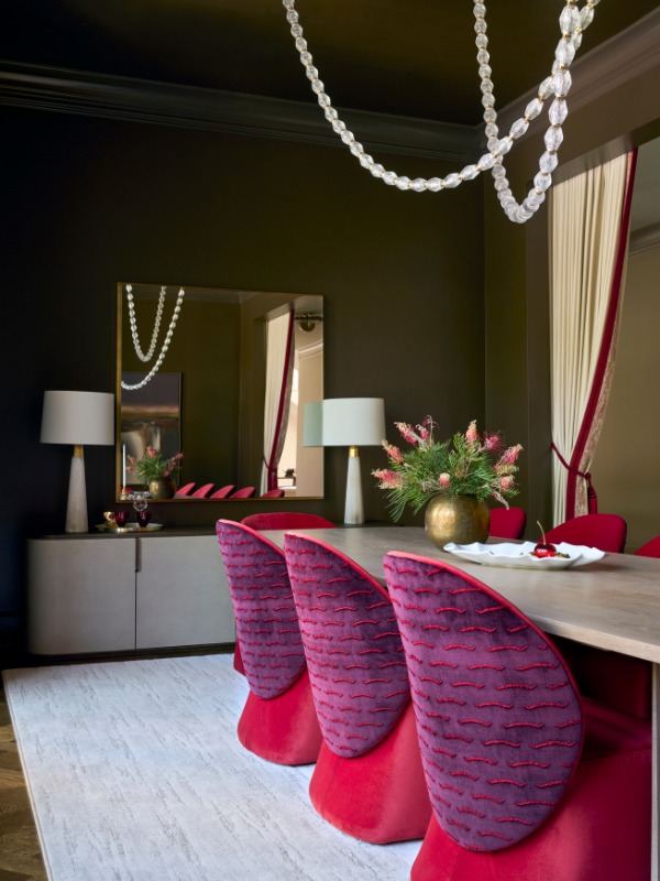 Moody dining room with deep olive walls, cherry-red and boysenberry chairs, a pale dining table, looping glass chandelier, and cream drapery trimmed in red.