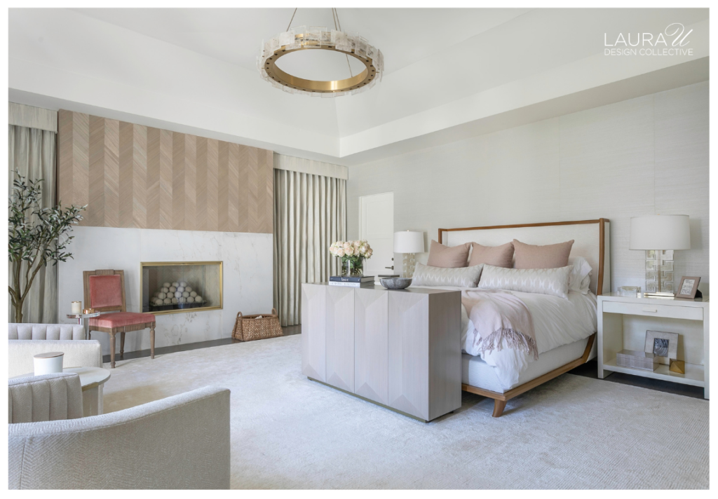 Soft neutral primary bedroom with a custom console at the foot of the bed, a marble fireplace, pale wood paneling, and blush accents.