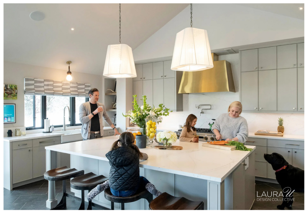 Family gathered around a large kitchen island beneath pendant lights, with gray cabinetry, a brass range hood, and natural light from multiple windows.