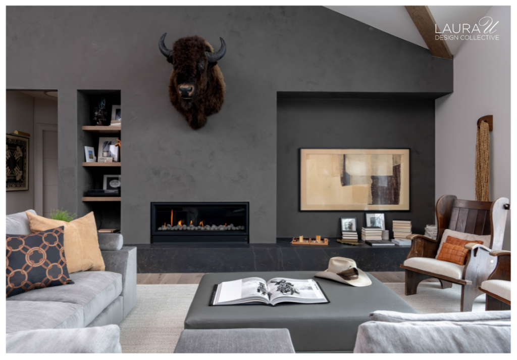 Modern living room with a dark gray fireplace wall, recessed shelving, stacked books, artwork, vintage wood chairs, and neutral upholstery.