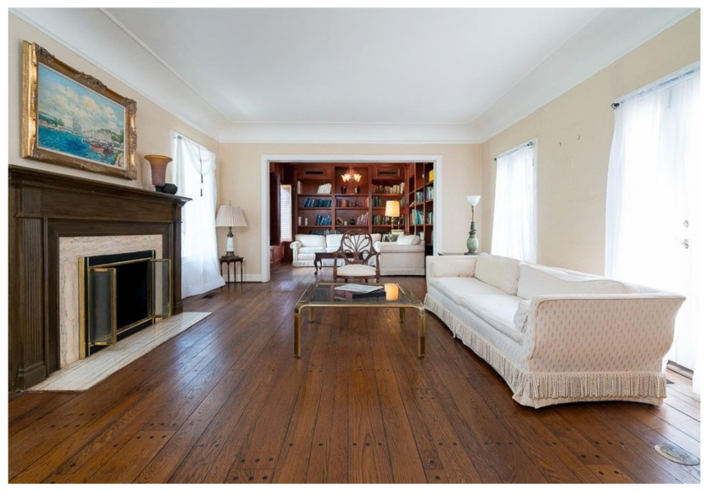Traditional living room before renovation with beige walls, dark wood floors, a fireplace, white sofa, and an adjoining wood-paneled library.