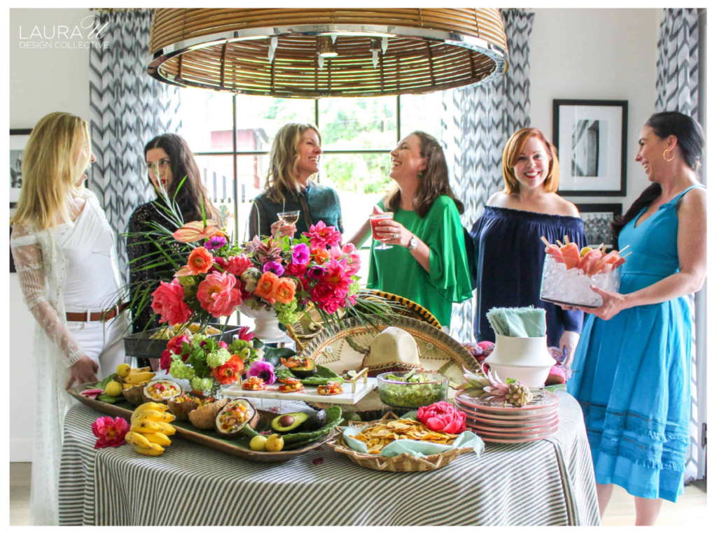 Guests gather around a flower-filled table with colorful seasonal arrangements, fresh food, and cocktails during a garden entertaining event.