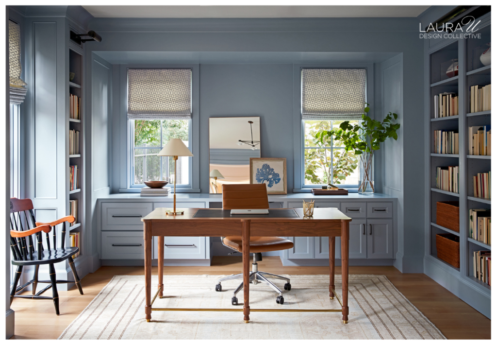 Blue home office with floor-to-ceiling built-in bookcases, closed lower cabinetry, a wood desk, and windows framed by Roman shades.